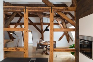 Un interior rústico con vigas de madera a la vista, un comedor con sillas y una mesa preparada para cuatro personas, y una cocina con electrodomésticos modernos.