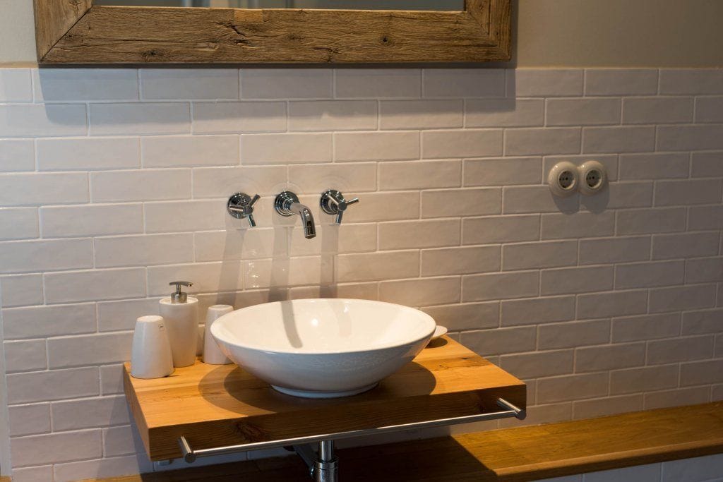 Ein Badezimmer mit einem weißen Keramikwaschbecken, zwei Wasserhähnen und einem Holzregal mit Toilettenartikeln.