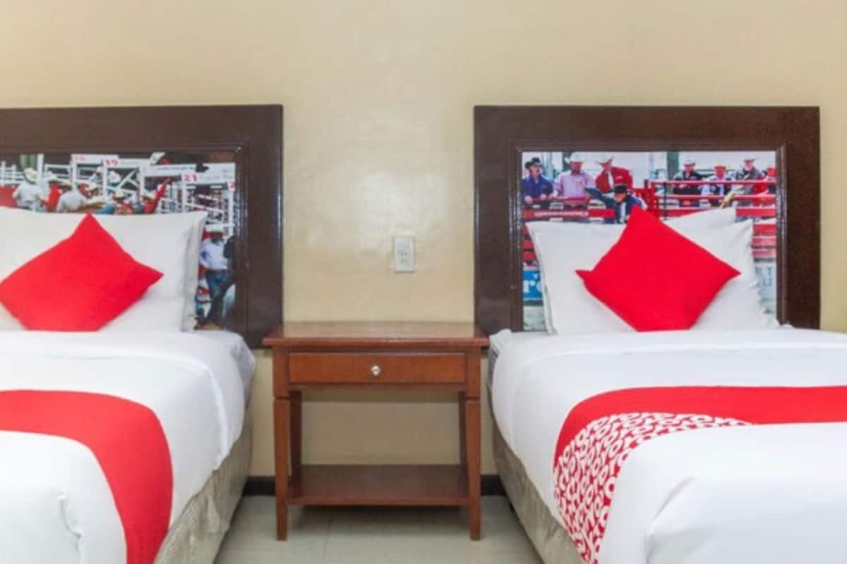 Ein Hotelzimmer mit zwei Betten, jedes mit roter und weißer Bettwäsche, einem kleinen Nachttisch aus Holz und einem an der Wand befestigten Fernseher, der ein Sportevent zeigt.