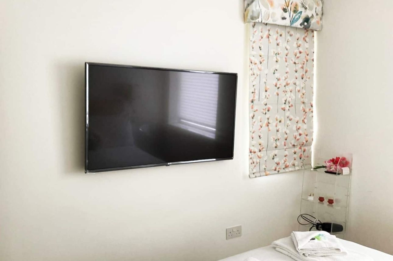 Een kamer met een wandgemonteerde flatscreen televisie, een raam met bloemen gordijnen en een klein kastje met toiletartikelen.