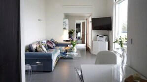 Ein modernes Wohnzimmer mit einer Couch, einem Couchtisch und einem Fernseher. Eine Tür führt in einen anderen Raum, und ein Fenster bietet einen Ausblick.