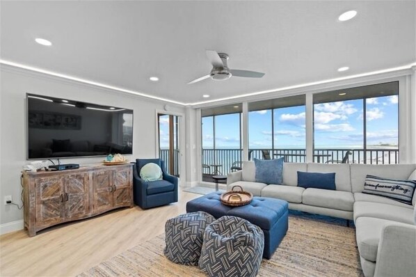 A spacious living room with a large TV, a ceiling fan, and a balcony with a view of the ocean.