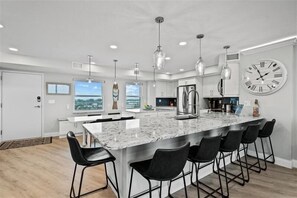 A modern kitchen with a large island, bar stools, pendant lights, and a large wall clock.