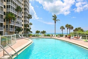 A swimming pool with lounge chairs and palm trees, overlooking a beach and a high-rise building.
