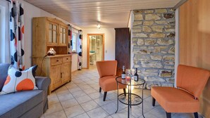 A cozy living room with a grey sofa, orange armchairs, a small round table with bottles, and a kitchen area with wooden cabinets and a stone wall.