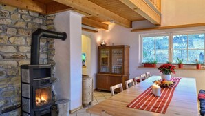 A cozy room with a stone wall, a fireplace, a wooden dining table with chairs, and a window with plants.