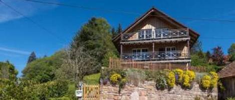 A wooden chalet with a balcony, surrounded by greenery and a stone wall.
