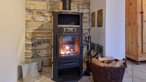 A wood-burning stove in a stone-walled room with a metal bucket and a wicker basket of firewood.