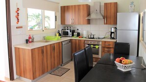 A modern kitchen with wooden cabinets, a dishwasher, a stove, and a dining table with a fruit basket. - Villa Ksenia (Paralimni)