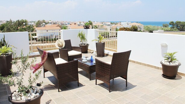 A terrace with wicker furniture, potted plants, and a view of the sea and town. - Villa Ksenia (Paralimni)