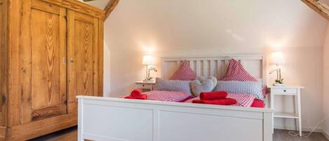Un dormitorio con una cama blanca, almohadillas a cuadros blancos y rojos, un armario de madera y dos mesitas de noche con lámparas.