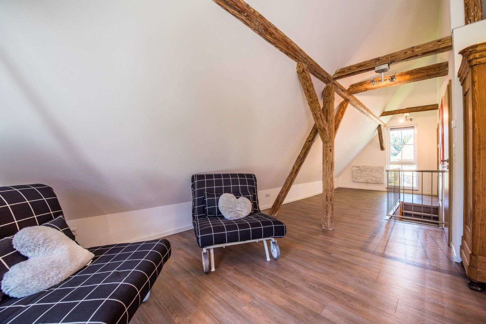 A loft-style room with exposed wooden beams, a sofa, a chair, and a view of a staircase leading to a balcony.