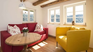 Ein Zimmer mit einem roten Sofa, einem gelben Sessel, einem Holztisch mit einer Vase und großen Fenstern mit Blick auf Bäume.
