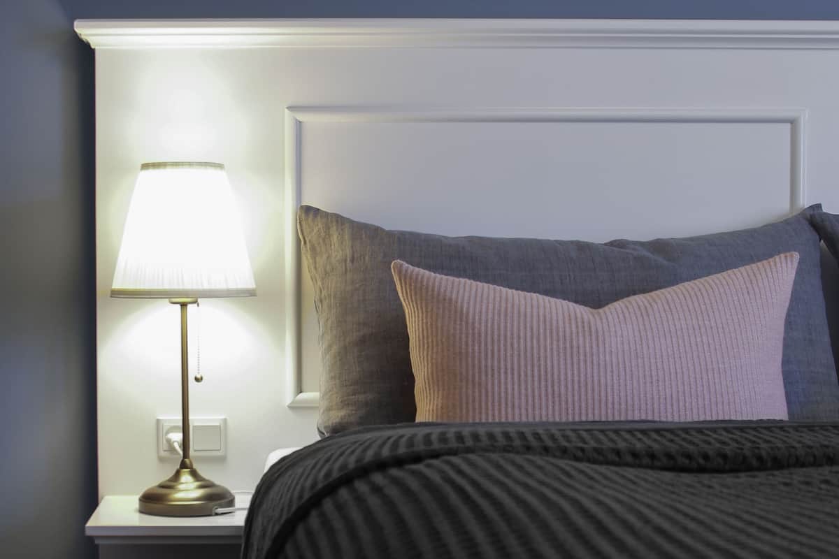 A neatly made bed with a dark-colored blanket and pillows, a bedside lamp, and a white nightstand.