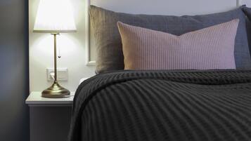 A neatly made bed with a dark-colored blanket and pillows, a bedside lamp, and a white nightstand.