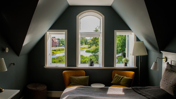 Une chambre avec un plafond en pente, deux fenêtres donnant sur la verdure, un lit recouvert d’une couvre-lit à motifs et une lampe de chevet.
