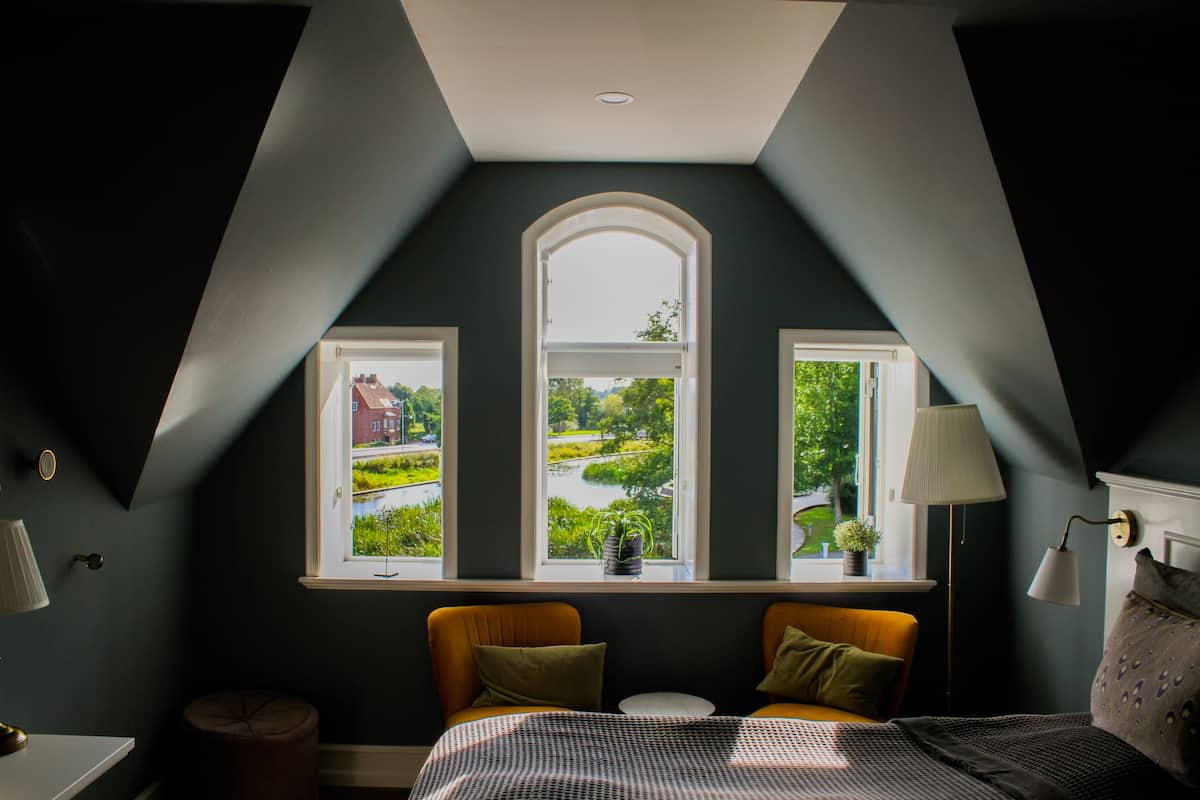 A bedroom with a sloped ceiling, two windows with views of greenery, a bed with a patterned bedspread, and a bedside lamp.