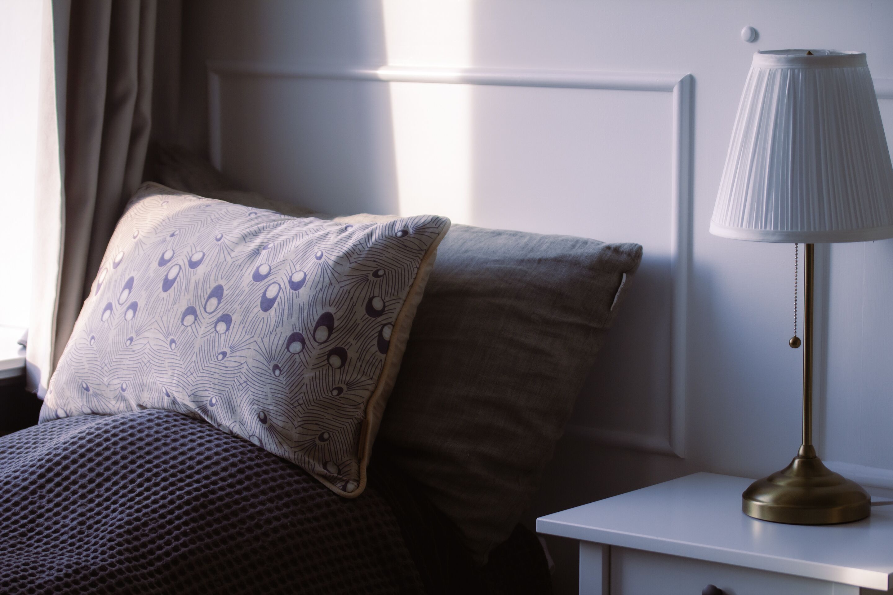 Una cama bien hecha con almohadas estampadas, una lámpara de noche y una ventana con cortinas.