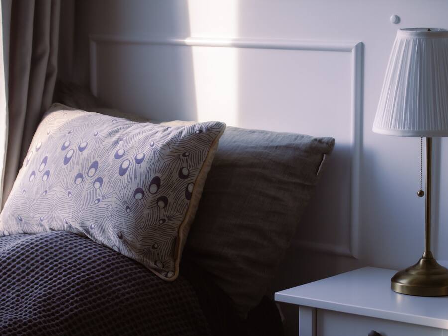 Una cama bien hecha con almohadas estampadas, una lámpara de noche y una ventana con cortinas.