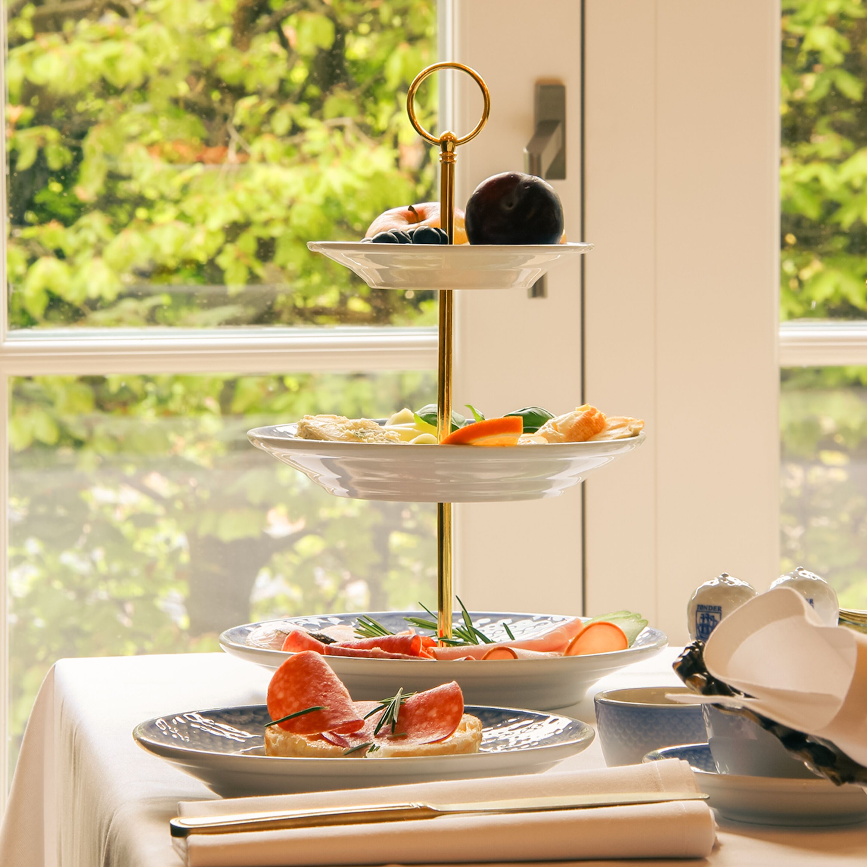 Un présentoir à trois niveaux, garni de diverses pâtisseries et de fruits, est placé sur une table d’où l’on peut admirer la verdure à travers une fenêtre.