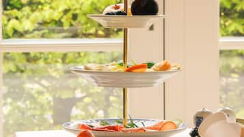Un soporte de tres niveles con diversos pasteles y frutas, sobre una mesa con vista a la vegetación a través de una ventana.