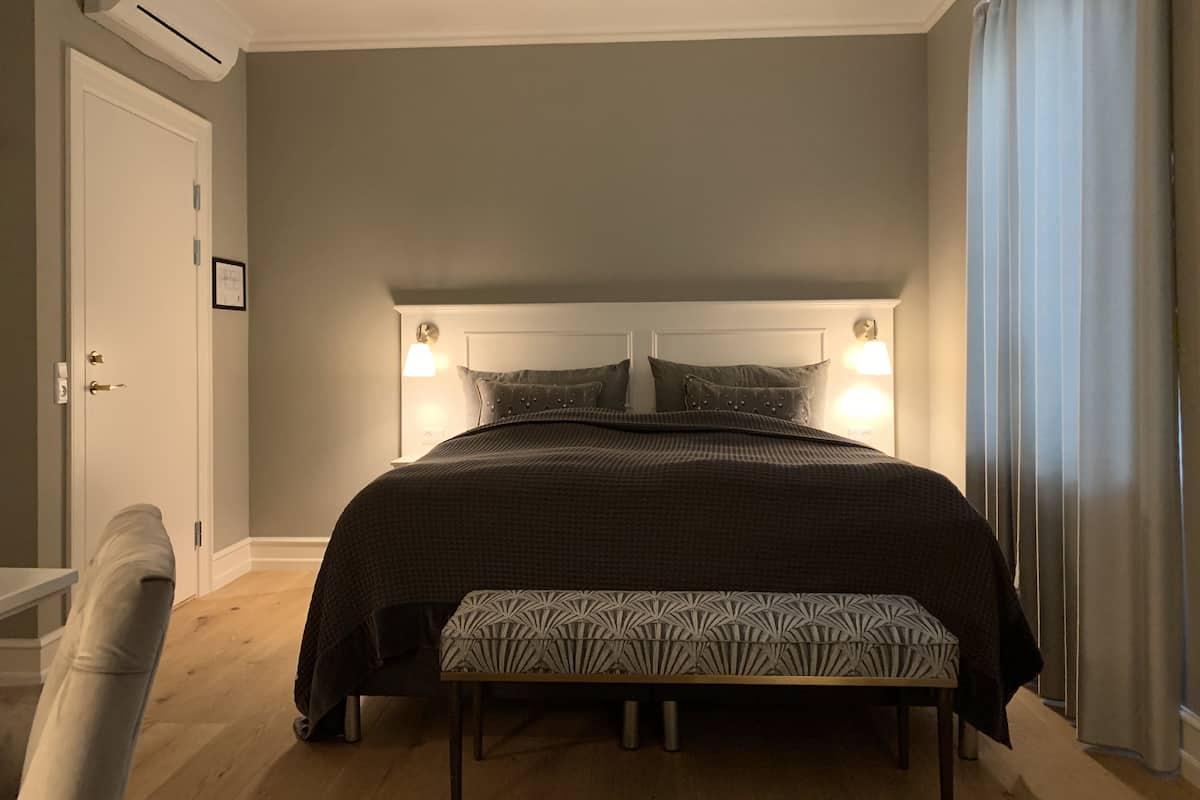 A bedroom with a bed, bedside lamps, a bench, a door, and an air conditioning unit.