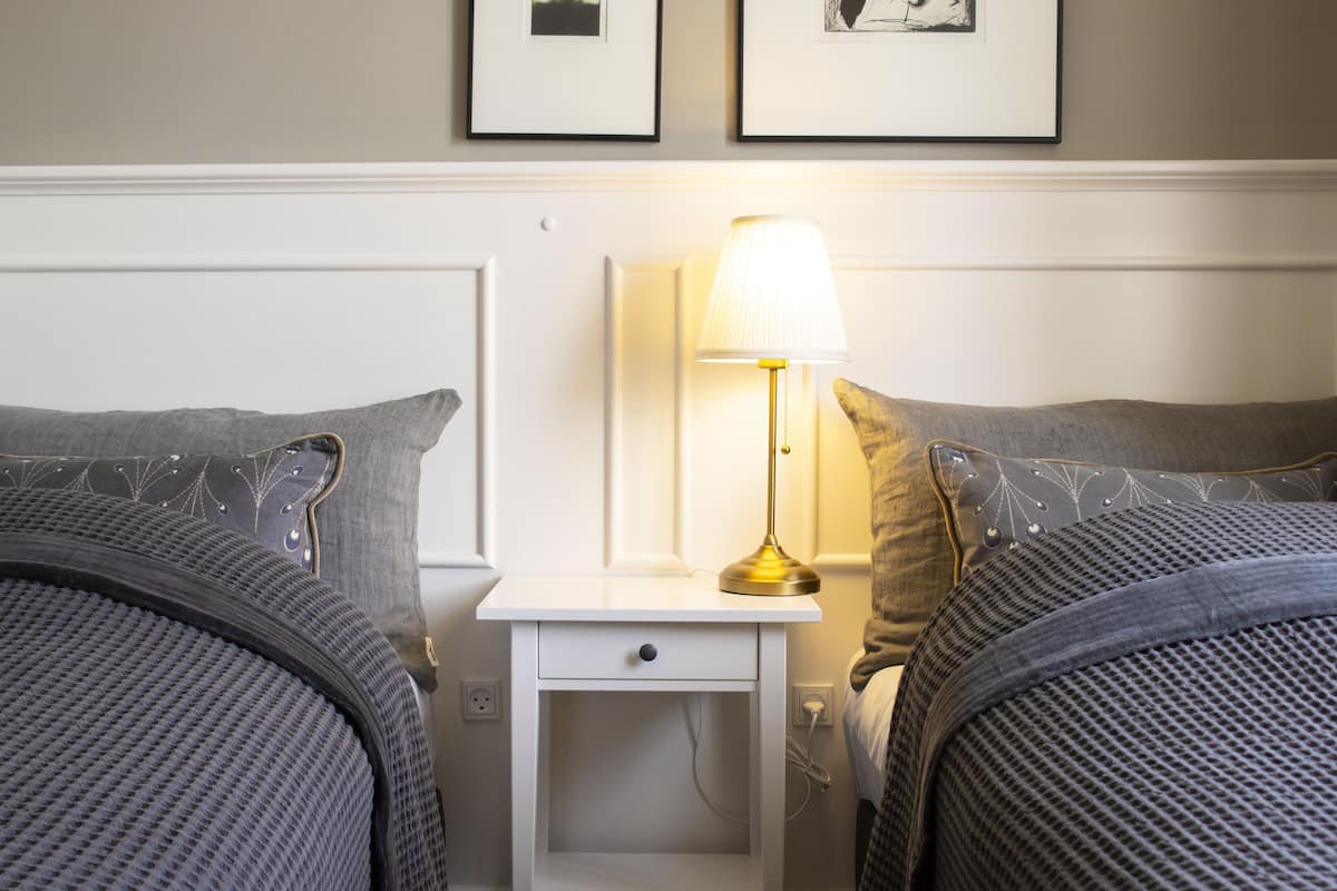 A neatly made twin bed with a grey headboard, a bedside table with a lamp, and a wall-mounted picture frame.