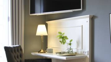 Ein Zimmer mit einem an der Wand befestigten Flachbildfernseher, einem Schreibtisch mit Lampe und einer Pflanze in einer Vase, einem schwarzen Polsterstuhl und einem Fenster mit Vorhängen.