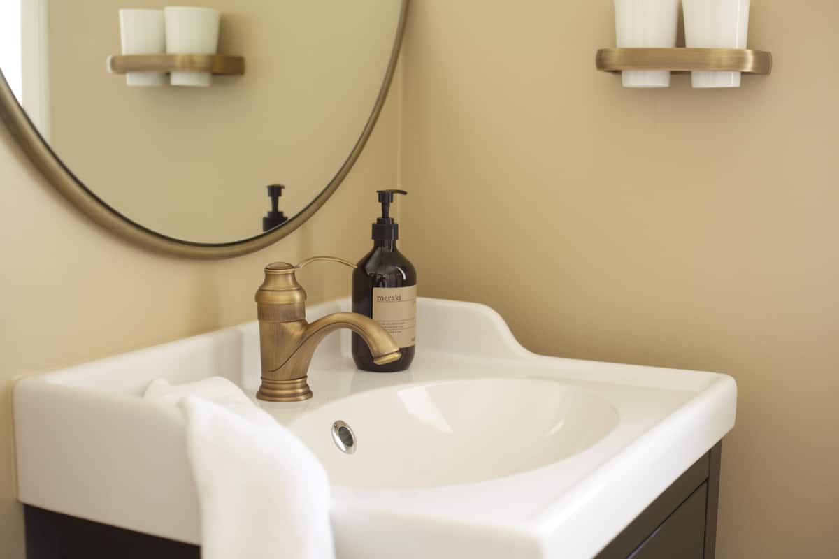 A bathroom with a white sink, a brass faucet, a bottle of Meraki soap, and a mirror.