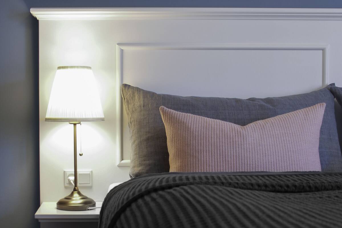 A neatly made bed with a dark-colored comforter and pillows, a bedside lamp, and a white headboard.