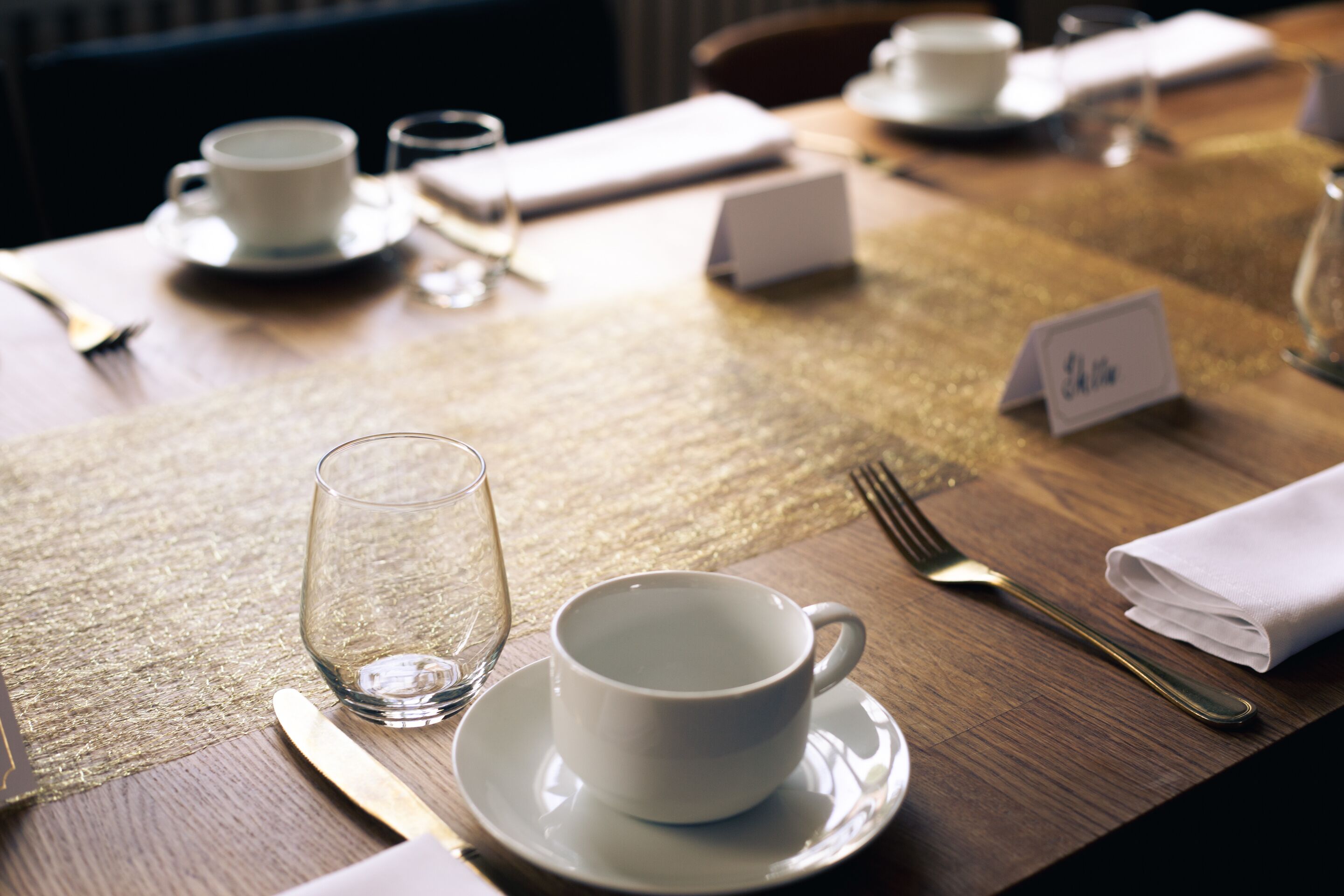 Una mesa de comedor dispuesta con una taza blanca, un platillo, un vaso y cubiertos.