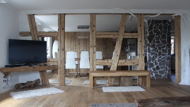 Un intérieur rustique avec des poutres apparentes, un téléviseur à écran plat, des chaises blanches et un mur en pierre.