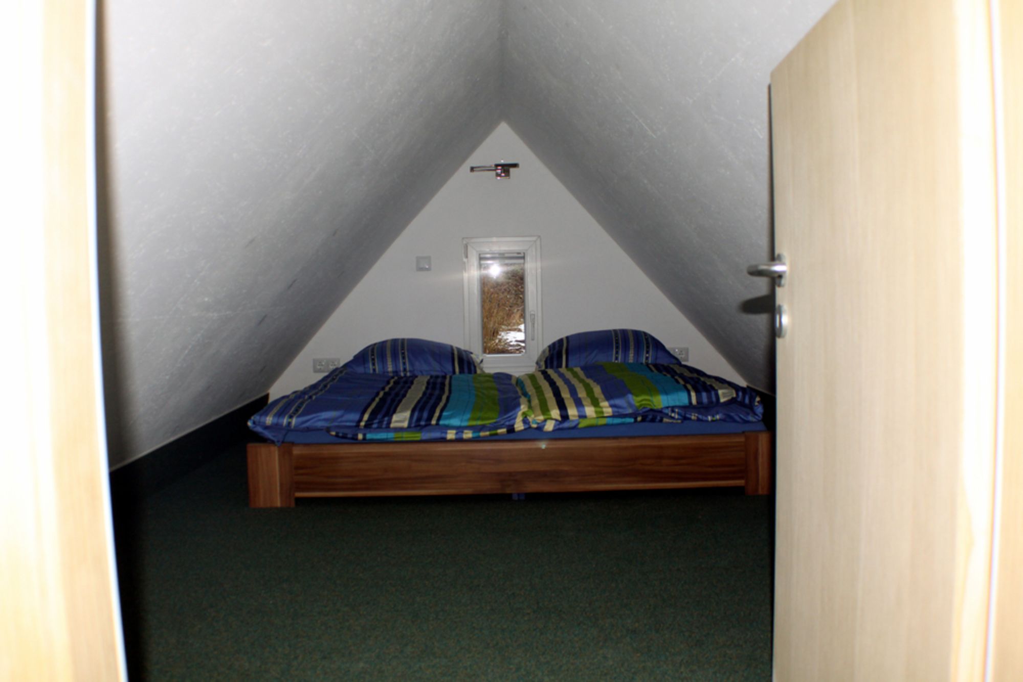 A small, triangular attic bedroom with a single bed, a window, and a door.