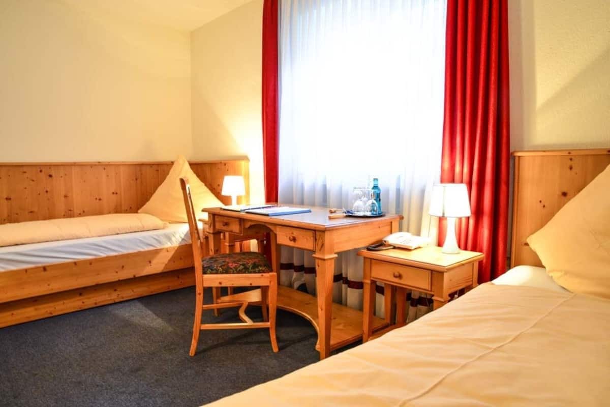 Habitación de hotel con una cama individual, un escritorio con silla, una mesita de noche con lámpara y una ventana con cortinas.