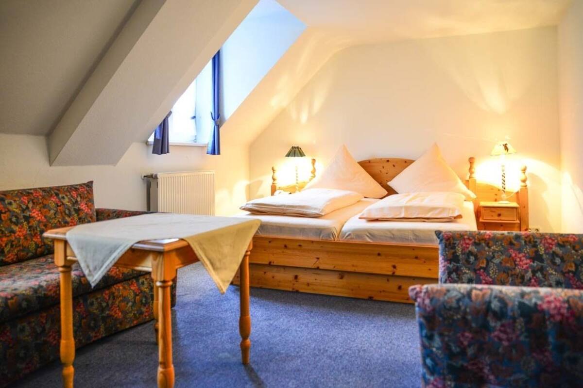 Un dormitorio acogedor con techo inclinado, una cama de madera, dos mesitas de noche con lámparas, una pequeña mesa con mantel y sillones estampados con motivos florales.