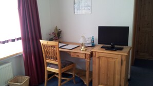 Een houten bureau met een computerscherm, een lamp en een stoel. Er is een raam met gordijnen en een aan de muur hangend schilderij.