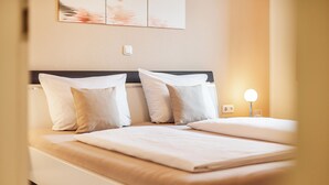Ein ordentlich bezogenes Doppelbett mit weißer Bettwäsche und zwei Kissen, ein Nachttisch mit Lampe sowie ein an der Wand befestigter Lichtschalter.