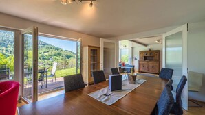 Une pièce spacieuse avec une grande table en bois, de nombreuses chaises et une vue sur un paysage verdoyant grâce à de grandes fenêtres.
