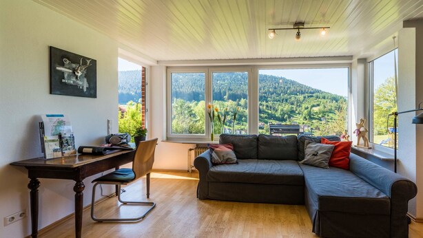 Un salon moderne avec un plancher en bois, un canapé gris foncé, un bureau en bois et une grande fenêtre donnant sur une colline boisée.