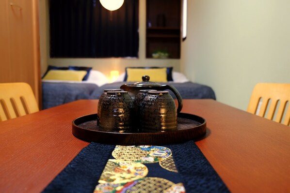 A wooden table with a tray holding two teapots, a blue table runner with a circular design, and a bed with a yellow pillow and a dark curtain in the background.