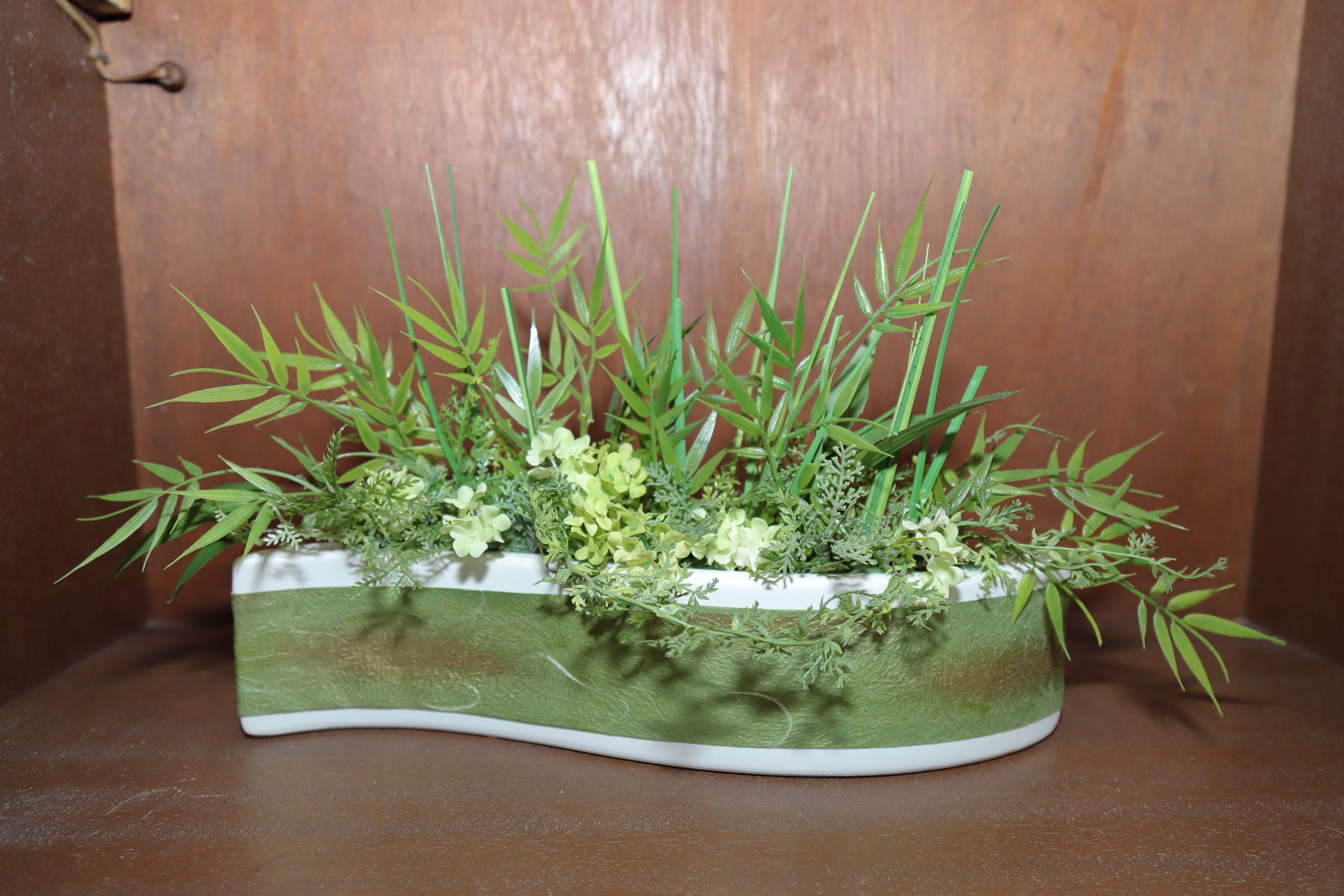 A rectangular planter with a green and white design, containing various green plants and small white flowers, placed on a brown surface against a wooden background.