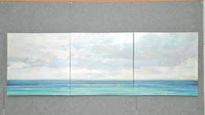 Una obra de arte grande de tres paneles que muestra un paisaje marino con aguas tranquilas y un cielo nublado, montada sobre una pared gris.
