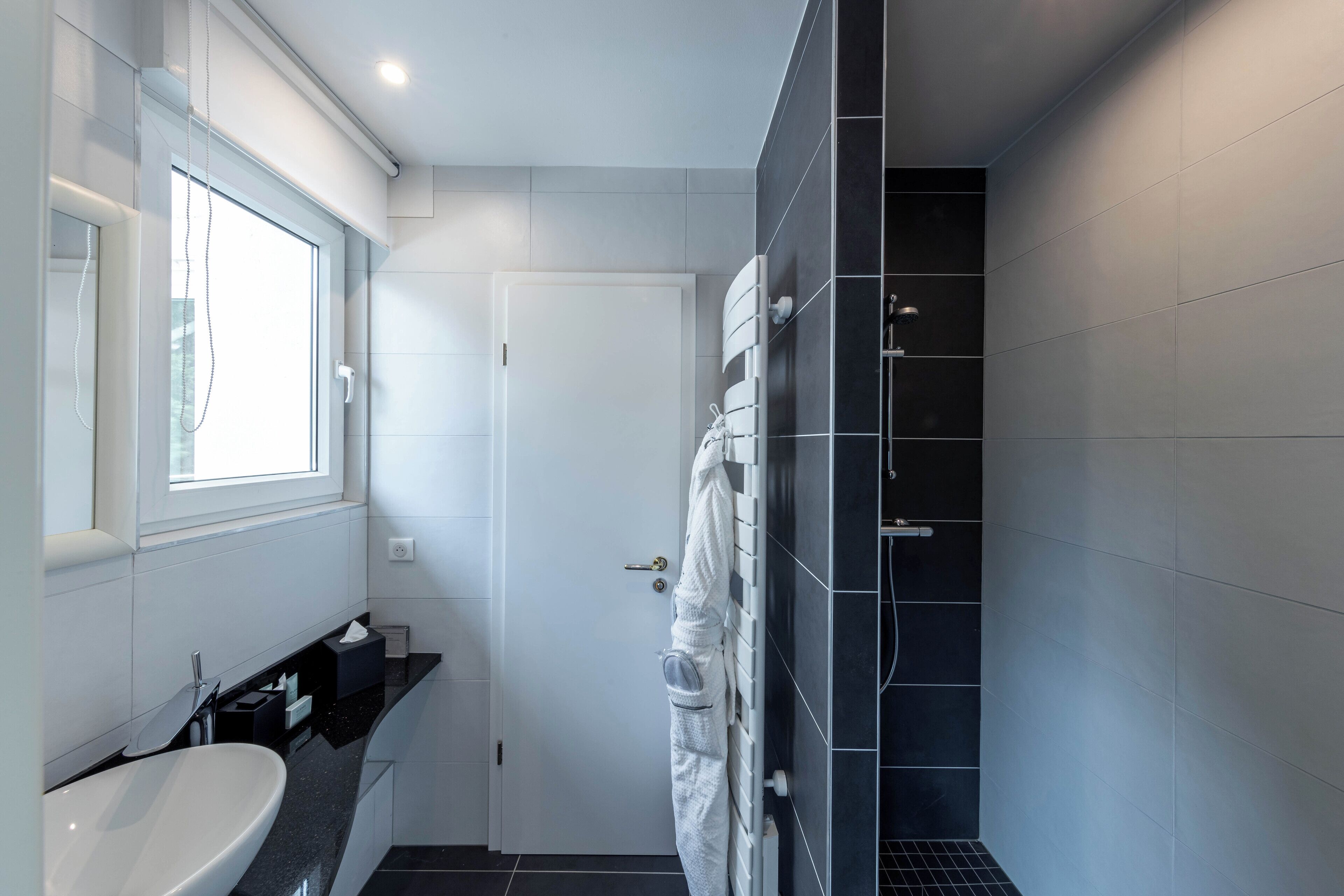 Een moderne badkamer met een witte wasbak, een douche met donkere tegels en een raam voor natuurlijk licht.