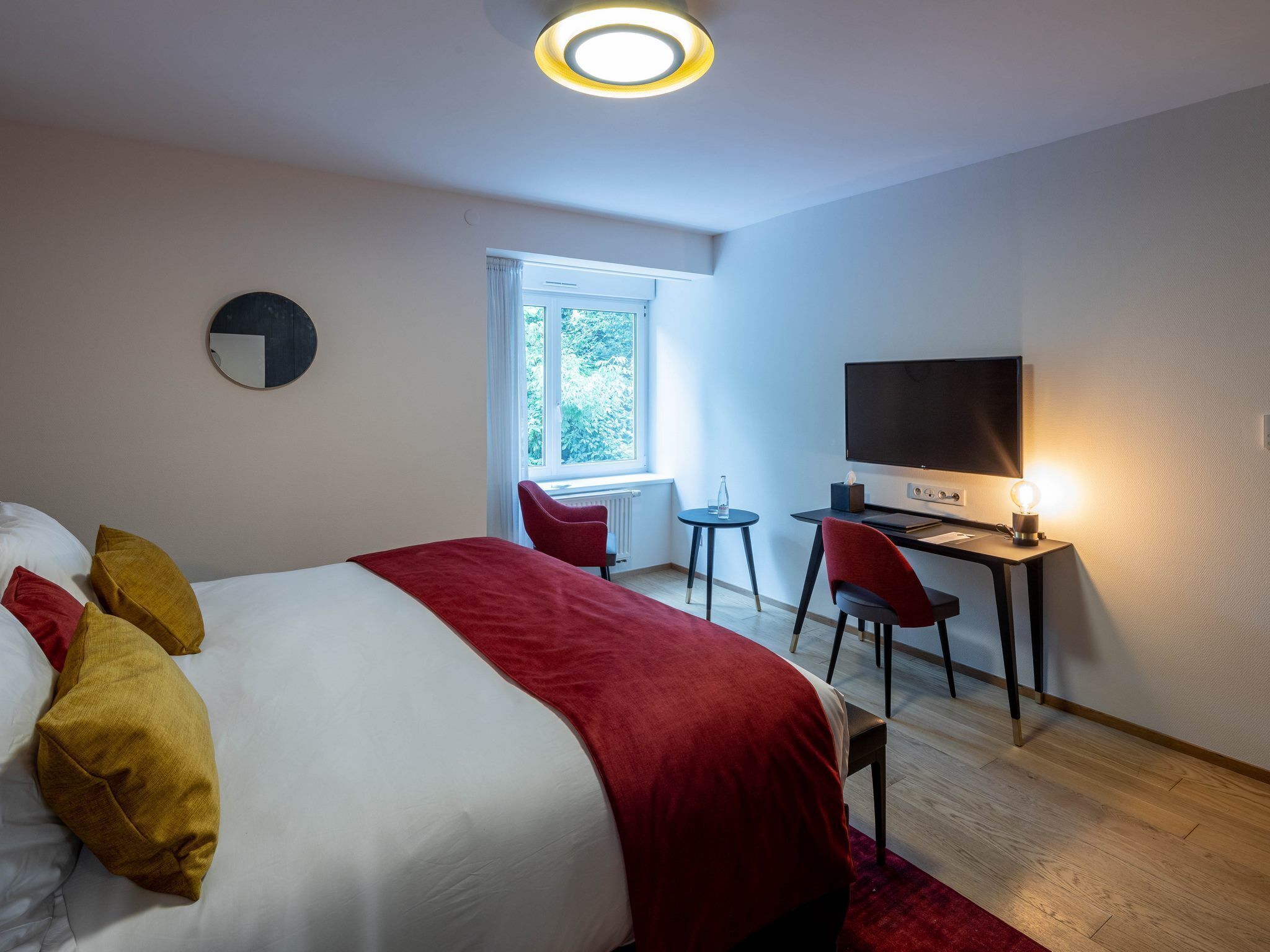 A modern hotel room with a large bed, a desk with a computer, a TV, and a window with a view of greenery.