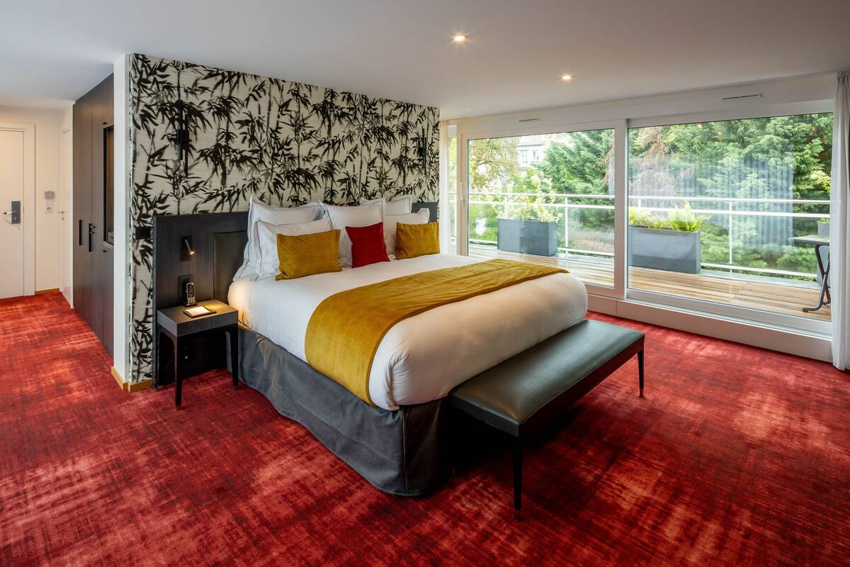 A modern bedroom with a large bed, a nightstand, a bench, and a view of a balcony with plants.