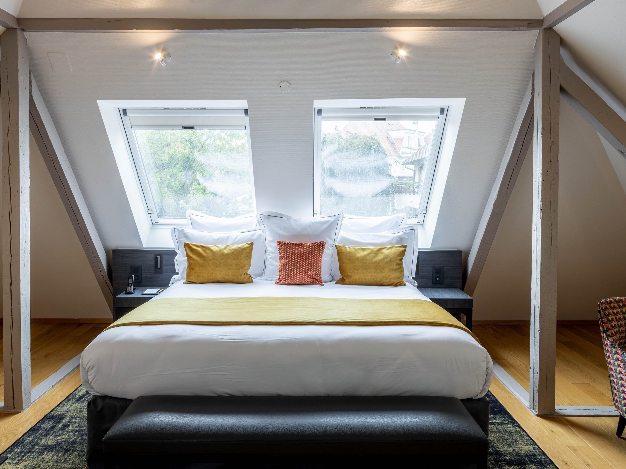 A bedroom with a large bed, two bedside tables, a bench, and two windows with a view of the outside.