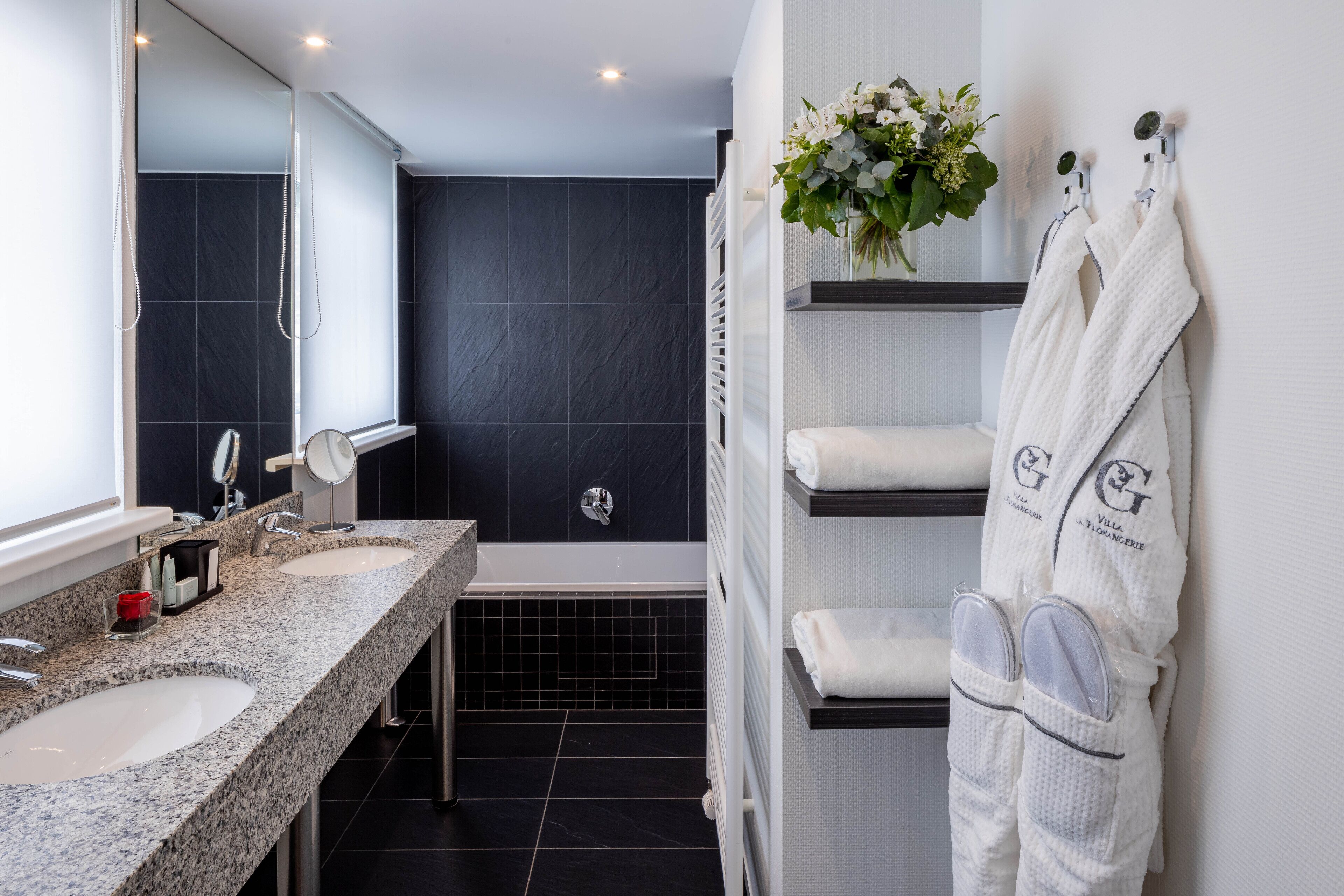 A modern bathroom with two sinks, a large mirror, dark tiled walls, and a shower area.