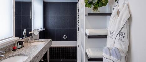 A modern bathroom with two sinks, a large mirror, dark tiled walls, and a shower area.