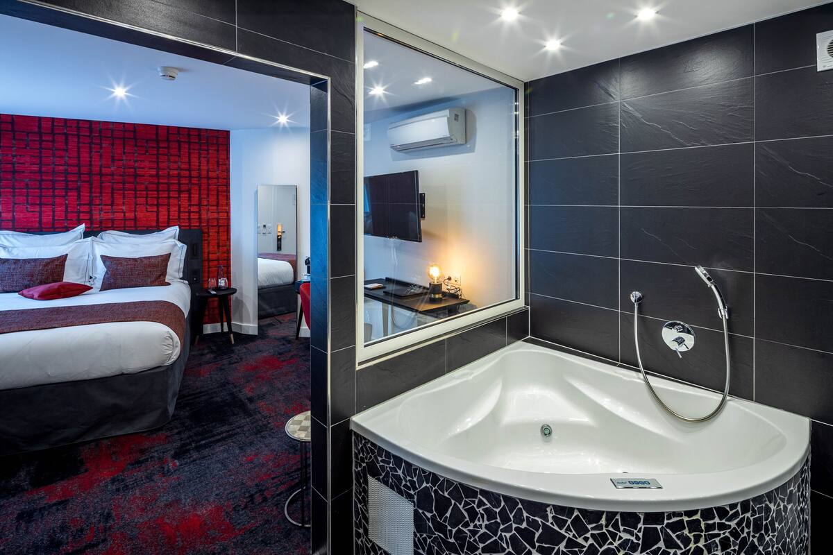 A modern hotel room with a large bed, a separate bathroom with a bathtub, and a red accent wall.