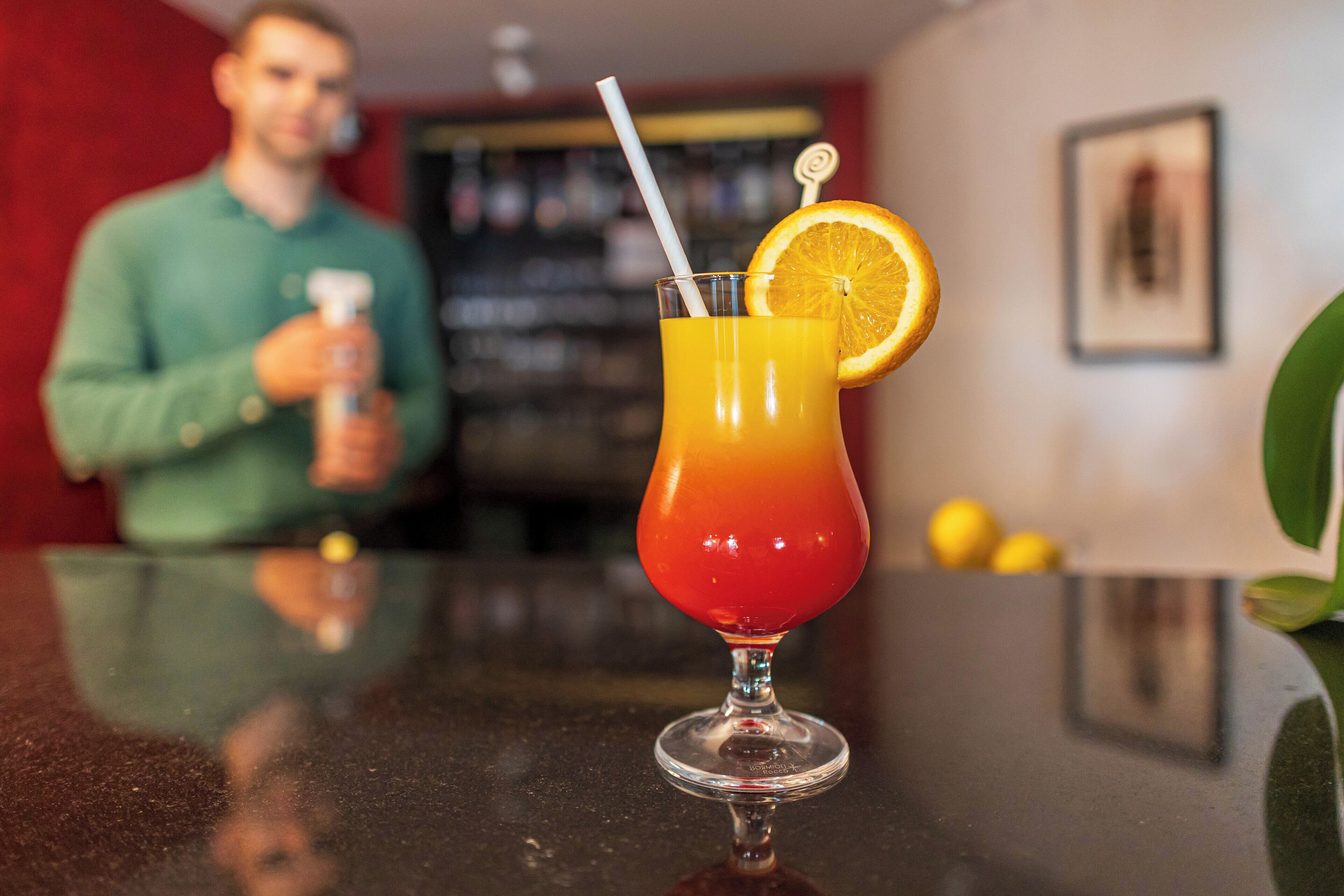 Una copa de cóctel con colores degradados, una pajita y una rodaja de limón sobre una superficie reflectante.